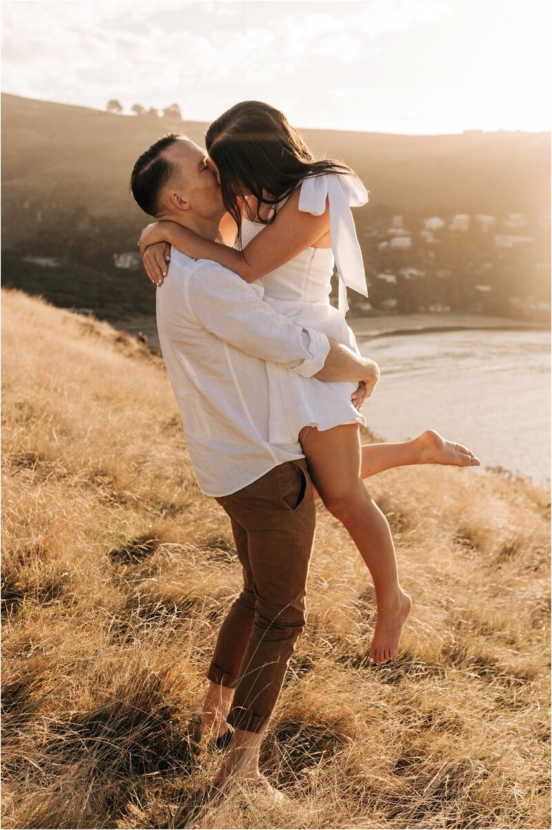 couple kissing sunset taylors mistake christchurch engagement white dress brown pants beach wedding photographer golden sun