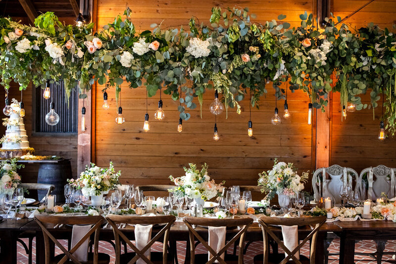 Wedding reception  with greenery, flowers and exposed bulbs hanging over head table.