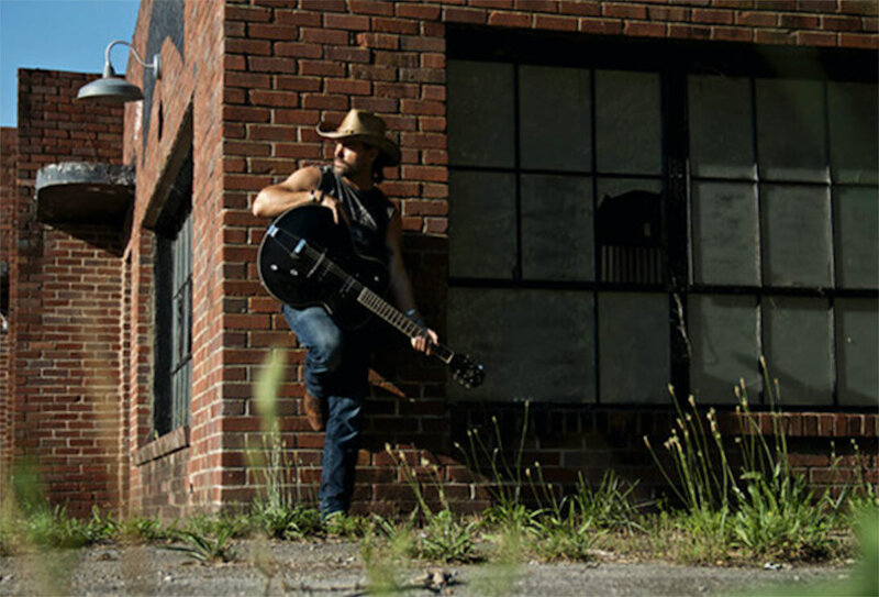 Country Music Photo Musician Billy Lord standing against brick building leaning on guitar he holds