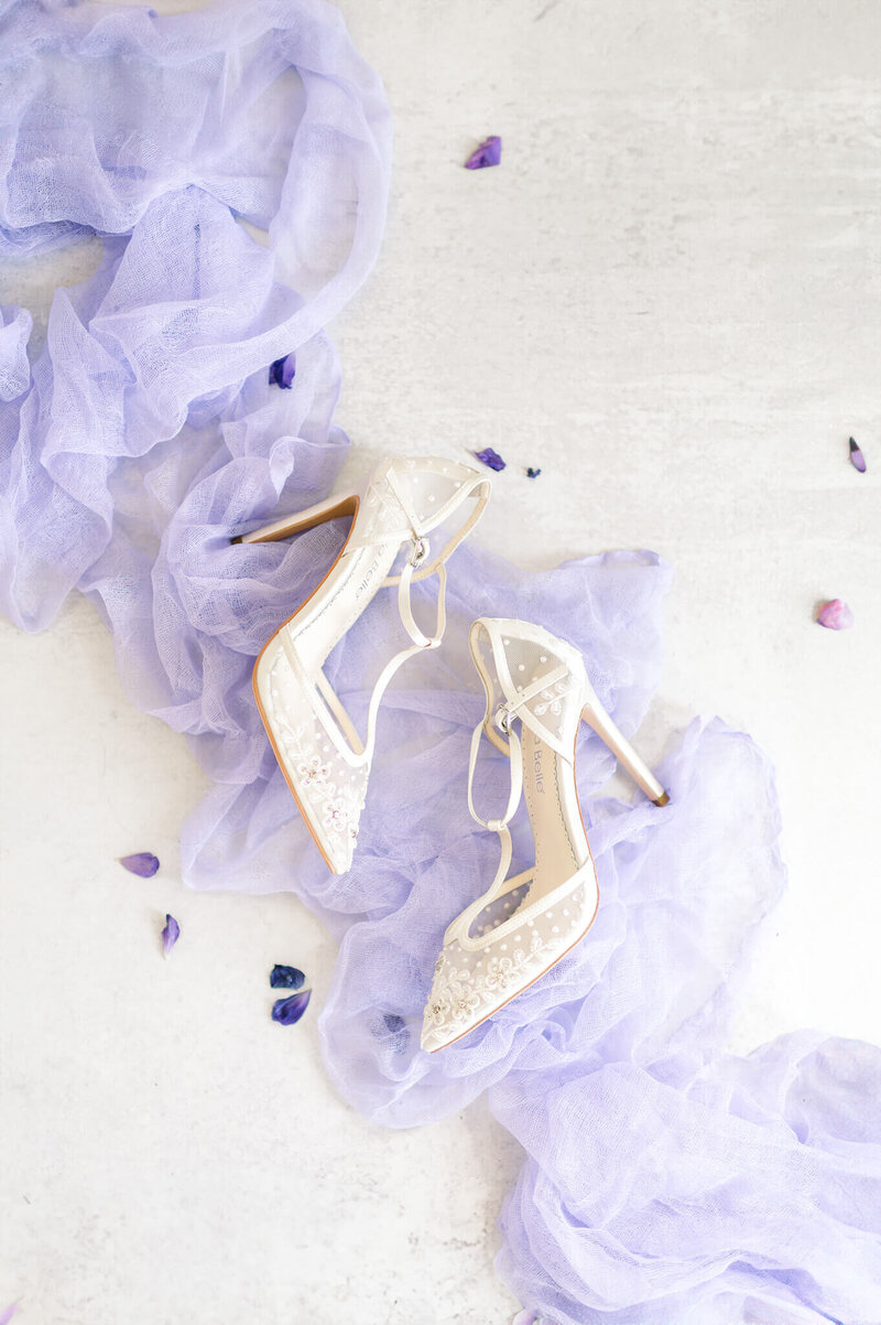 white bridal heels sitting on soft purple fabric with purple flower petals scattered around