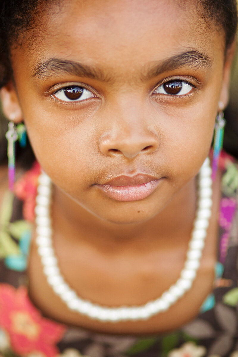 Close up shot of a beautiful young girl with piercing brown eyes. You can see that she has on a pretty, colorful dress and a white necklace.