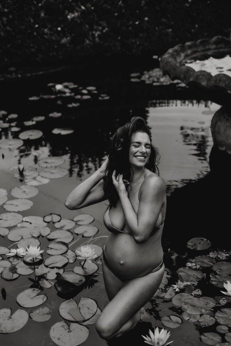 Creative maternity photography with a pregnant woman in a lily-filled pond, laughing and holding her hair back.