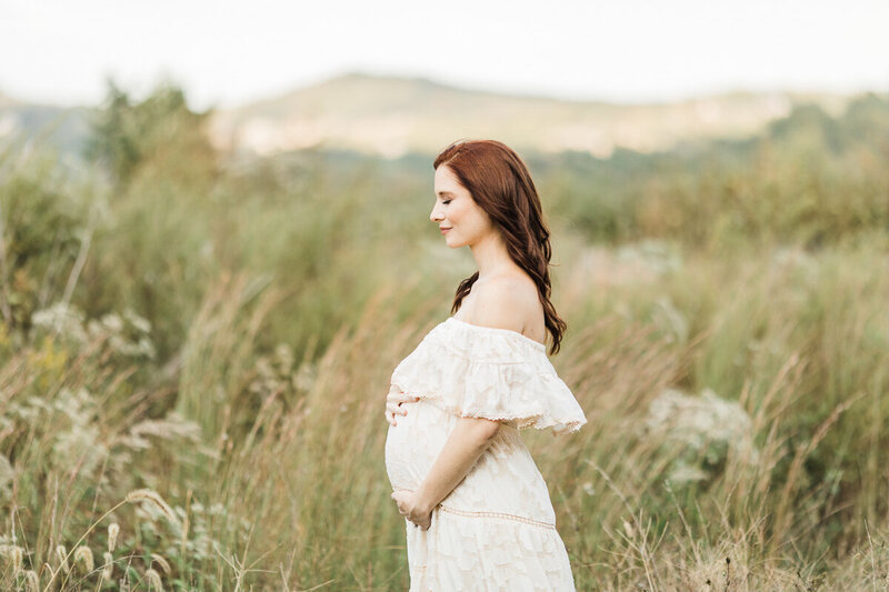 Outdoor maternity photo of redheaded mom in a white dress on scenic hillside