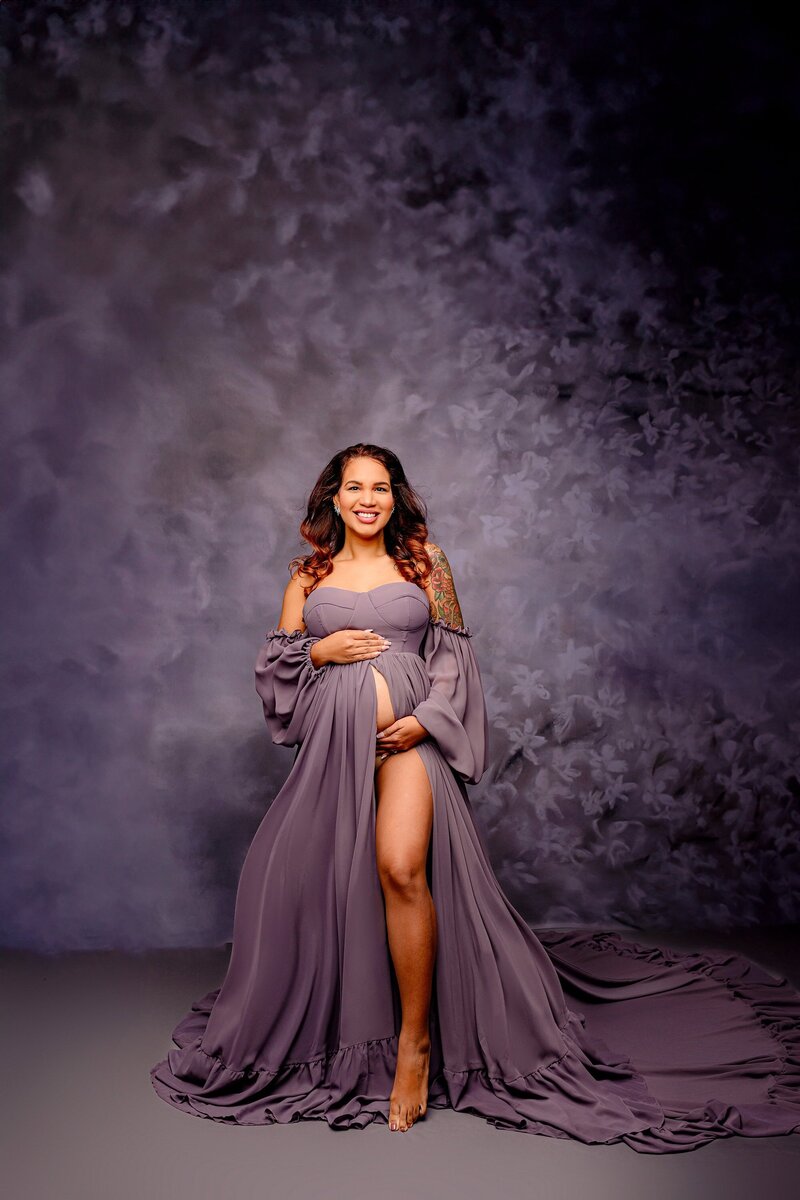 Expectant Charlotte mother in a Dusty purple dress with one leg out and both hands on her belly on a Studio background