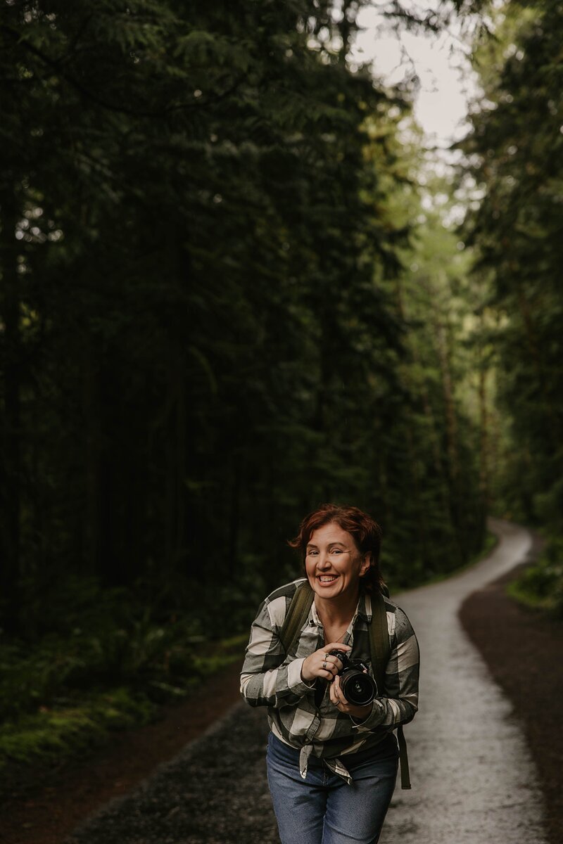 A smiling woman holding a camera walks on a forest path surrounded by tall, green trees, capturing behind the scenes at a creative photography retreat.