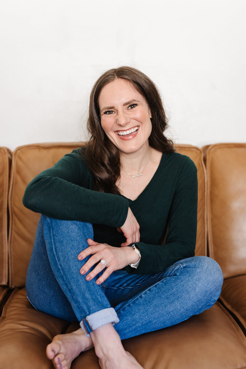 woman sitting on brown couch with leg up smiling at the camera