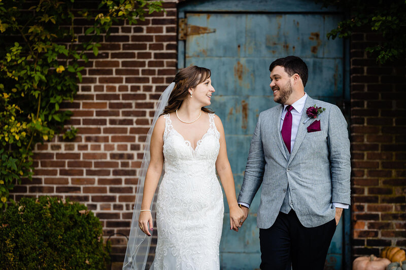 A newlywed couple holding hands and smiling at each other, with the bride in a lace gown and the groom in a grey suit, standing in front of a rustic blue door and brick wall covered in ivy.