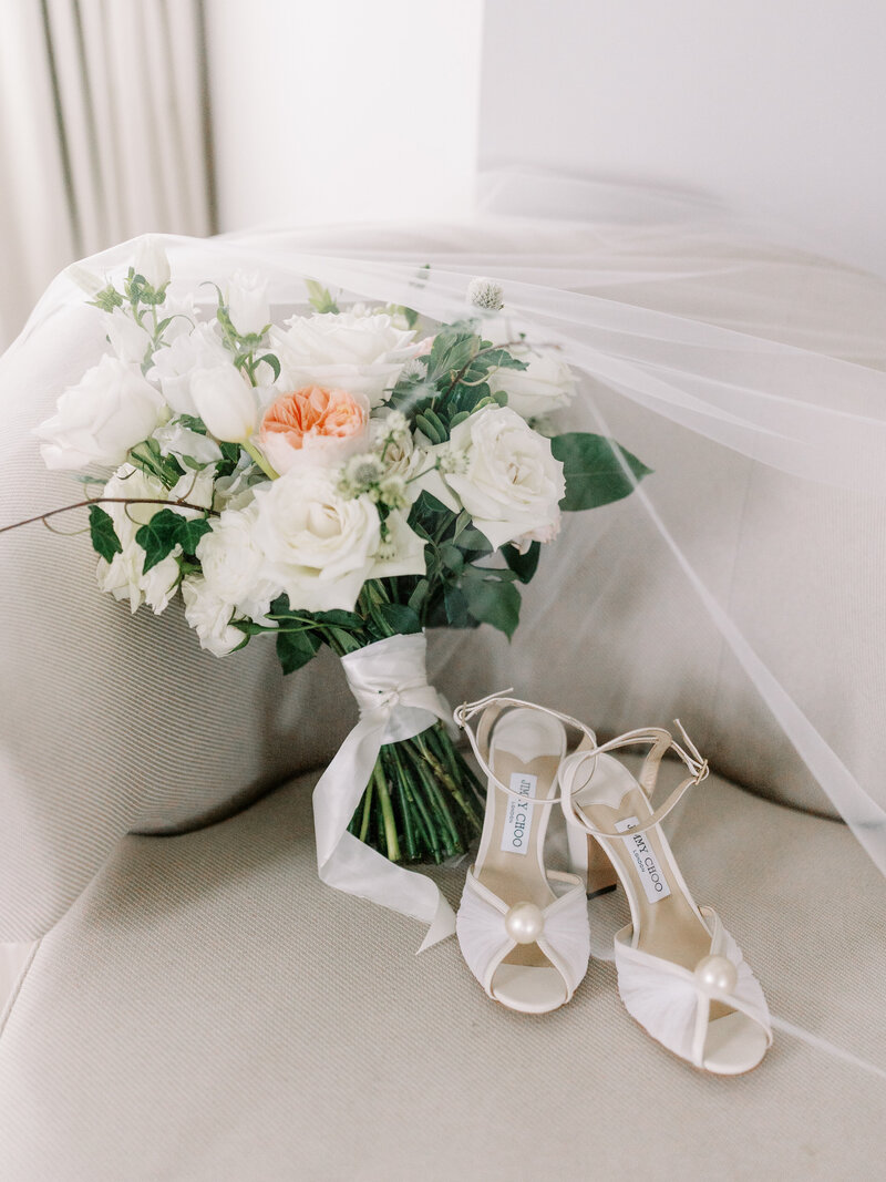 Bridal bouquet of white flowers laying on a couch next to bride's wedding shoes