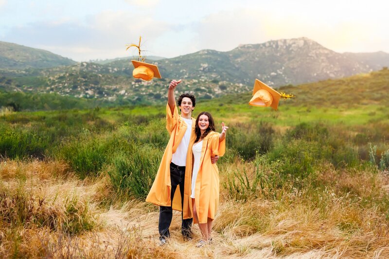 Two Temecula Valley high school seniors standing in a field with mountain views while throwing caps toward the camera