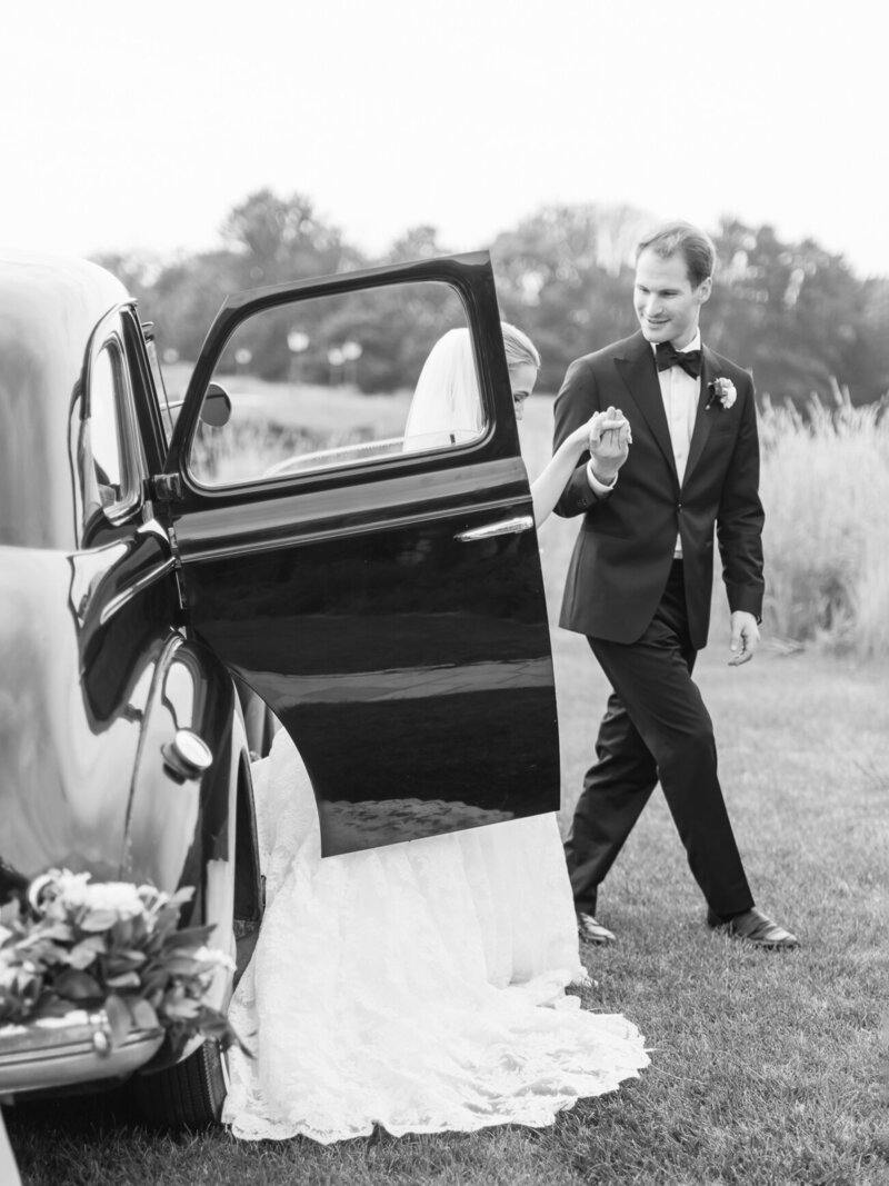 A black and white photo of a groom in a tuxedo helping his bride get out of a vintage car.