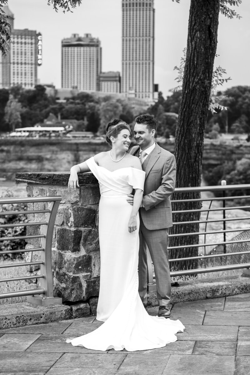 Bride and groom in elegant black-and-white portrait standing by stone railing with city skyline in background, Niagara Falls NY, photographed by wedding photographer Niko Coric - Lumen Clarity Media