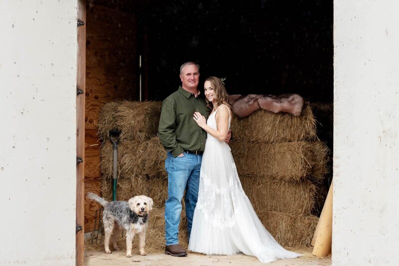 a bride and groom stand in front of bales of hay with their dog looking on