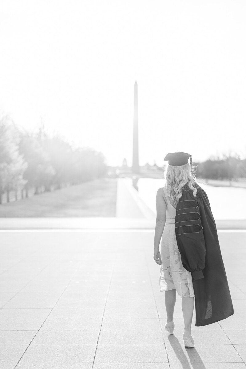 girl wearing graduation cap holding gown while walking towards Washington Monument during senior pictures