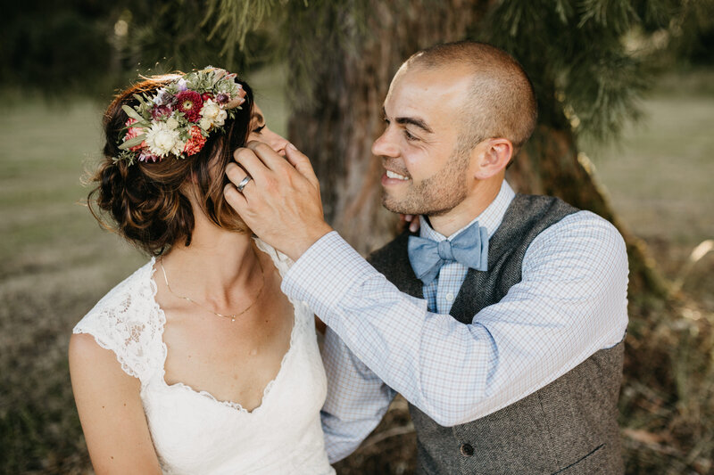 Bride wears a flower crown and is smiling at groom who is touching her face.