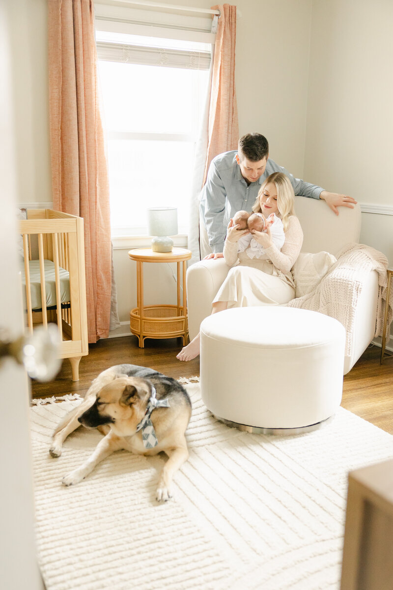 mother holds twin newborns while sitting in a cream glider as her husband leans beside her in the nursery with their dog, Indianapolis newborn photographer