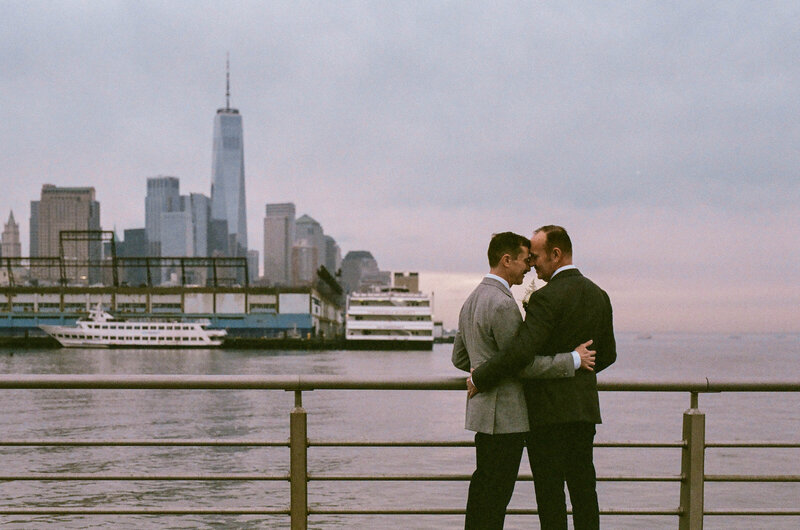 A couple with their arms around each other as they stand overlooking a city skyline across the water.
