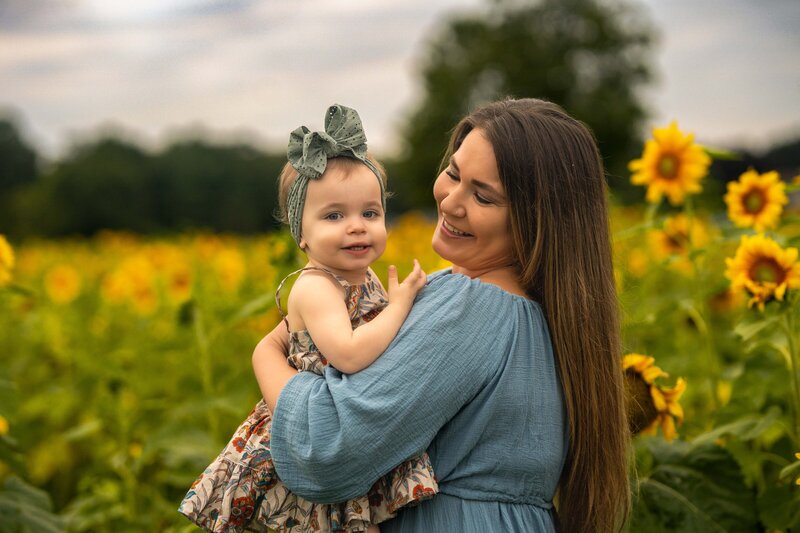 mom in a blue dress holding her toddler as toddler smiles at camera and mom smiles at toddler in medina sunflower field ohio