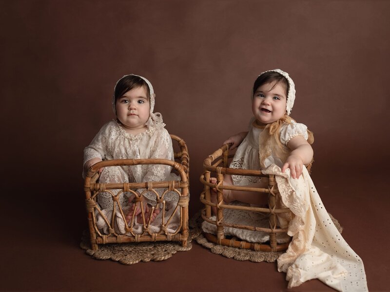 Twin toddler girls sit in wicker baskets in matching white dresses and bonnets posed by a Lafayette Baby Milestone Photographer