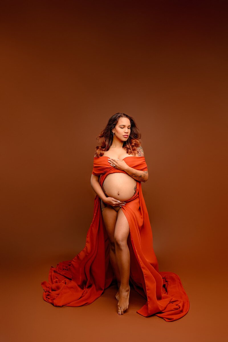 charlotte area mom is looking down with both hands on her pregnant belly with her body wrapped in a rust colored chiffon wrap