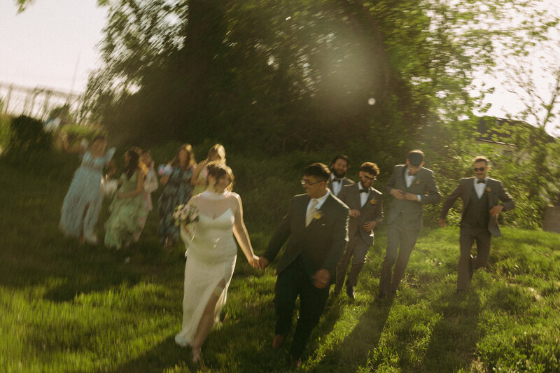 A groom and bride celebrate their union by joining arms and walking down the aisle after their ceremony. Her dress trails behind and shines in the sun's glow