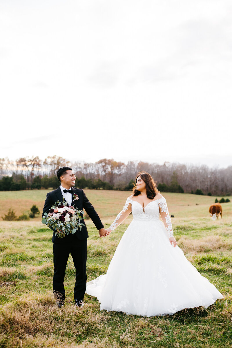 Shenandoah wedding venue at a horse farm with bride and groom in the horses pasture as they hold hands and smile while looking at one another with a winter tree line in the distance