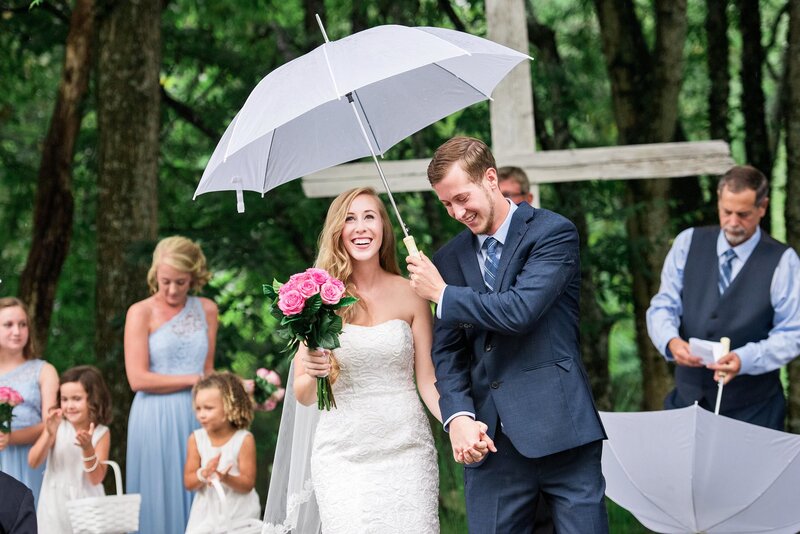 Groom holding white umbrella over him and his wife during outdoor rainy ceremony
