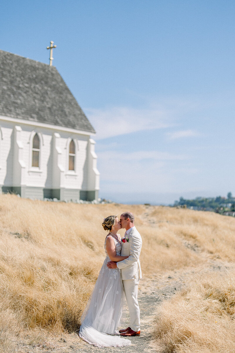 Bride and groom kiss in golden California grass behind a white church, photographed in editorial wedding style near San Francisco.