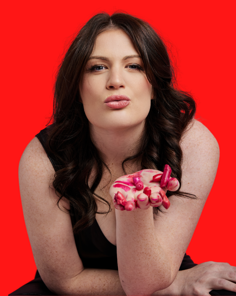 In front of a red background, Emily appears to be blowing a kiss to the camera but she is holding up her hand which is covered in broken pieces of lipstick in shades of red and pink