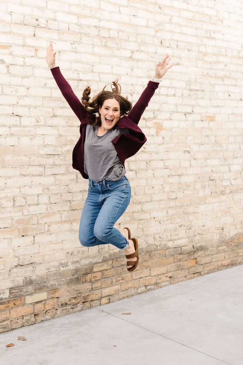 woman jumping in the air with arms raised in front of a brick wall