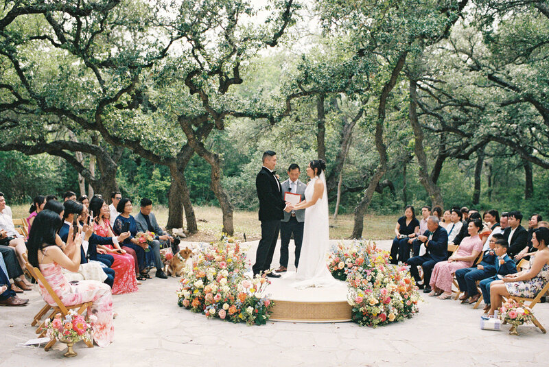 A couple stands on a flower-adorned platform exchanging vows in a serene outdoor wedding ceremony. Guests are seated on both sides, surrounded by lush trees.