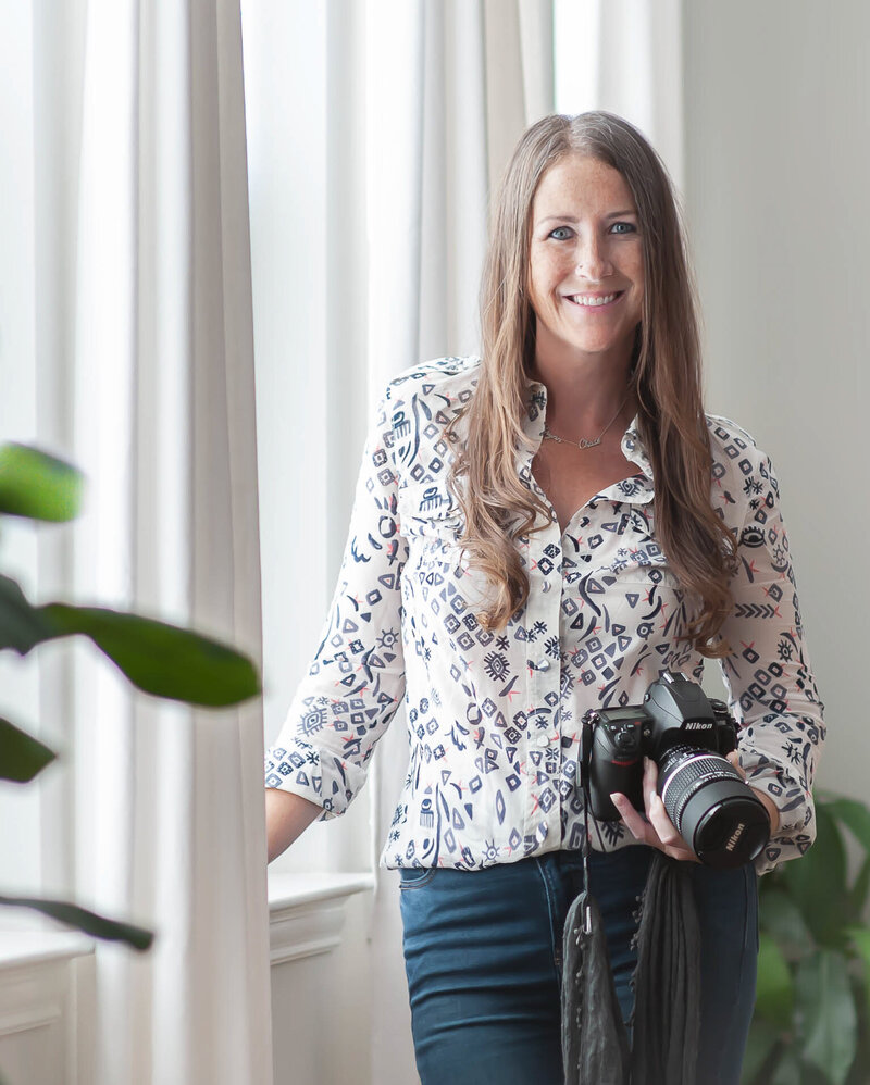 Professional Photographer Allison Amores holding camera near a window with white curtains and plants in the foreground and background