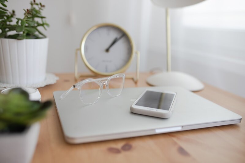 Photo shows a small brass clock, glasses in clear frames, a cell phone lying on a laptop