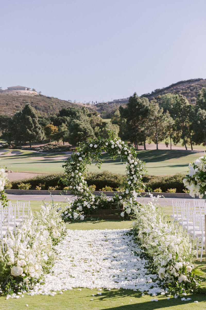 Outdoor wedding ceremony space, the aisle is liked with lots of fluffy white flowers, and completely covered in white petals. At the end there is a cicular floral archway