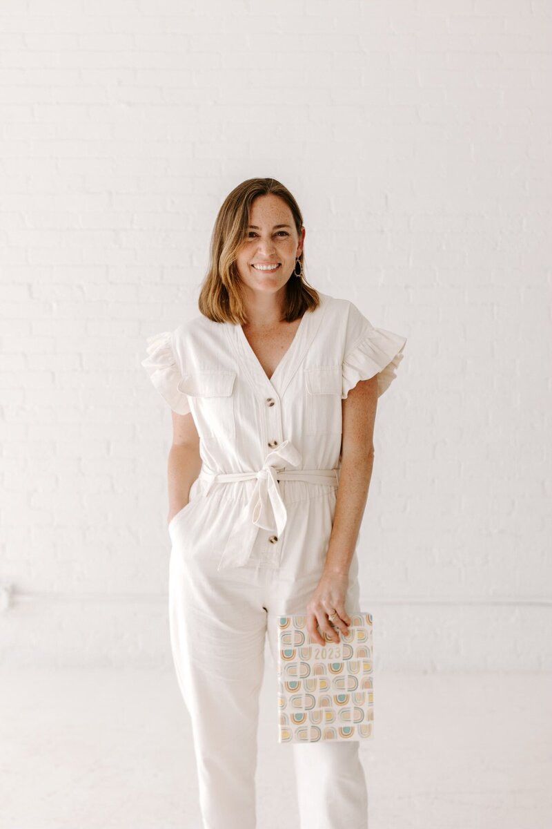 Chicago wedding planner smiles at camera while holding notebook. She wears a white linen romper and her hand is in her pocket.