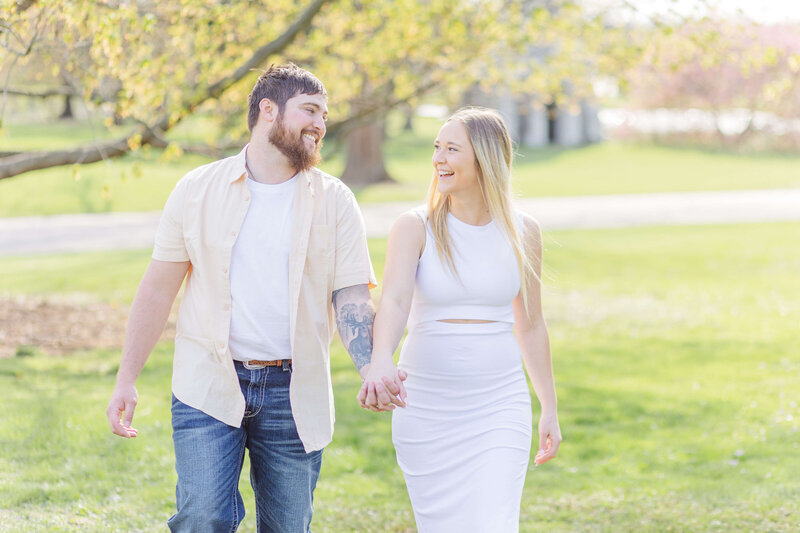 A man and woman are walking in a park holding hands and laughing. It's springtime and the trees have yellow flowers. She is wearing a white dress and he is wearing a yellow shirt.
