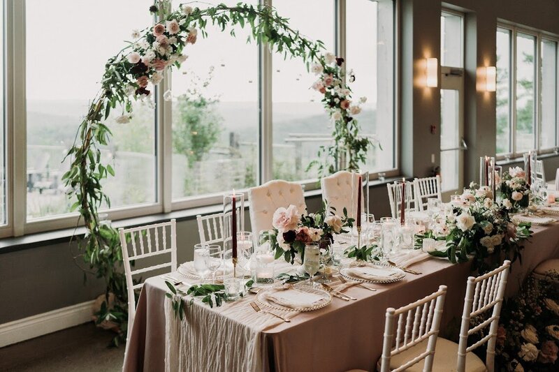 A large floral moongate backdrop sits behind the dusty rose head table filled with burgundy tapered candles and flowers at this romantic wedding at Le Belvedere venue in Wakefield Quebec.