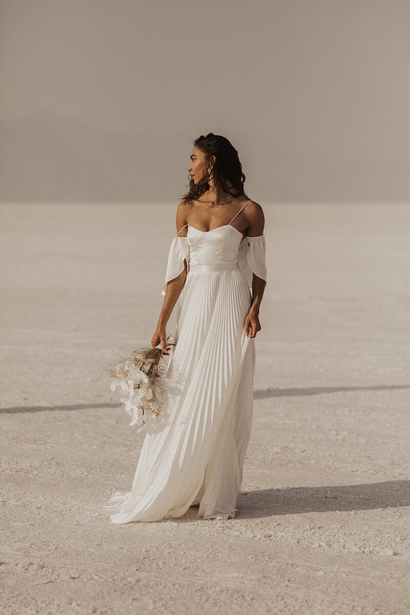 A woman in an elegant white dress holding a bouquet on a vast salt flat.