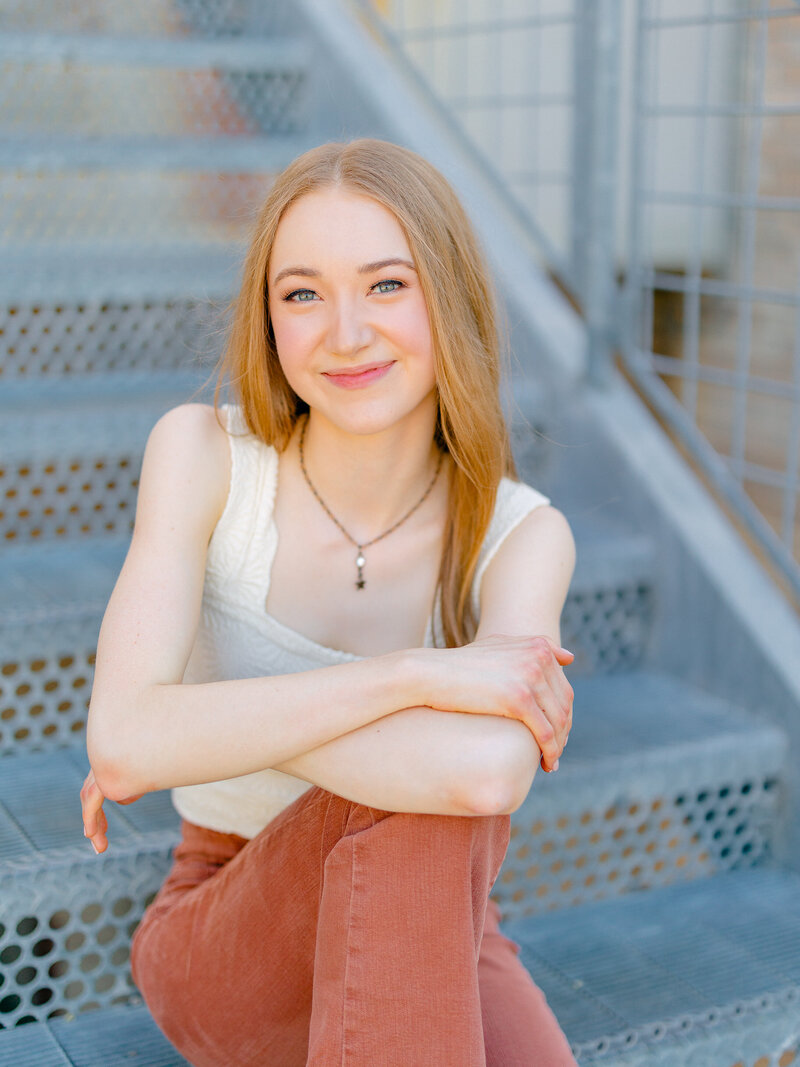 High school senior in rust long pants and white top sitting on blue stairs