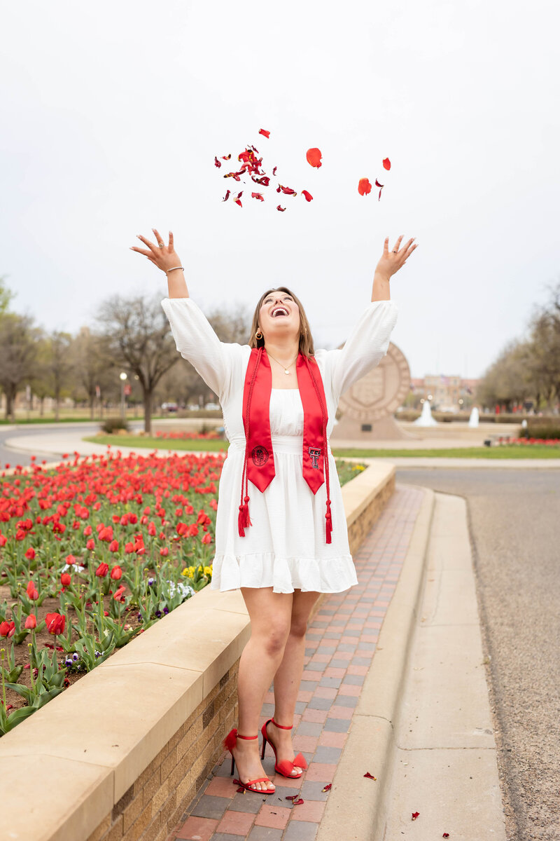 woman graduate is wearing a white dress with red heals and stole with cords and is smiling as she is tossing  red flower petals at the entrance of Texas Tech University Campus