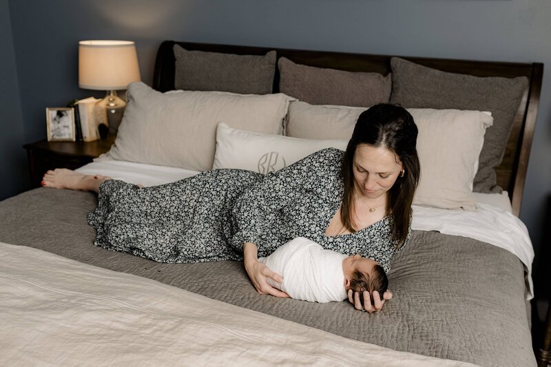 Mother lays on bed looking down towards her newborn son the baby is dressed and a cream wrap and mom wears a flower dress and their Charlotte Area home during their lifestyle newborn session
