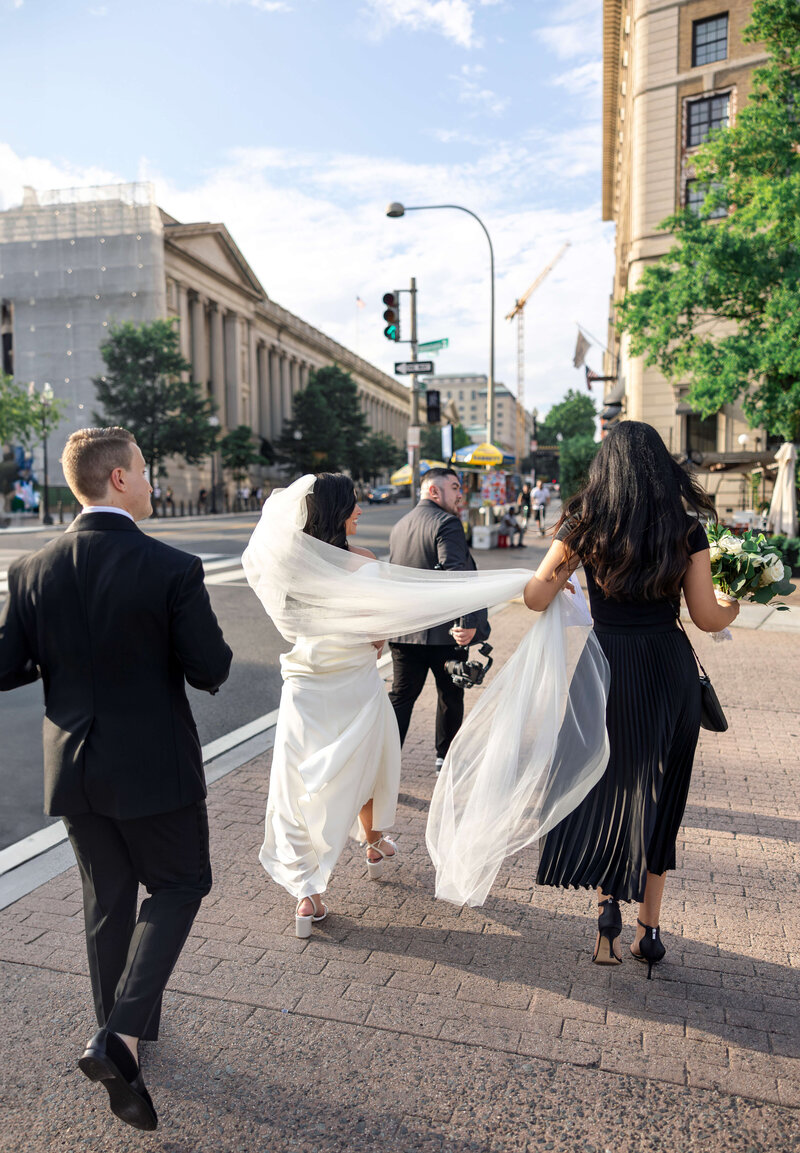 A bride in a white dress walks outdoors with her bridal party. Two people assist with her veil while another person accompanies them. Theyre on a city street, surrounded by buildings and trees under a partly cloudy sky.