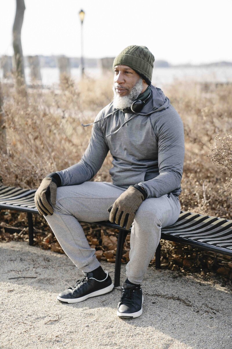 A masculine presenting person of color with a curly white beard sits on a bench, looking past the viewer with a gentle smile. They are wearing a gray hoodie, green beanie hat, and headphones around their neck. Their hands rest gently in their lap.