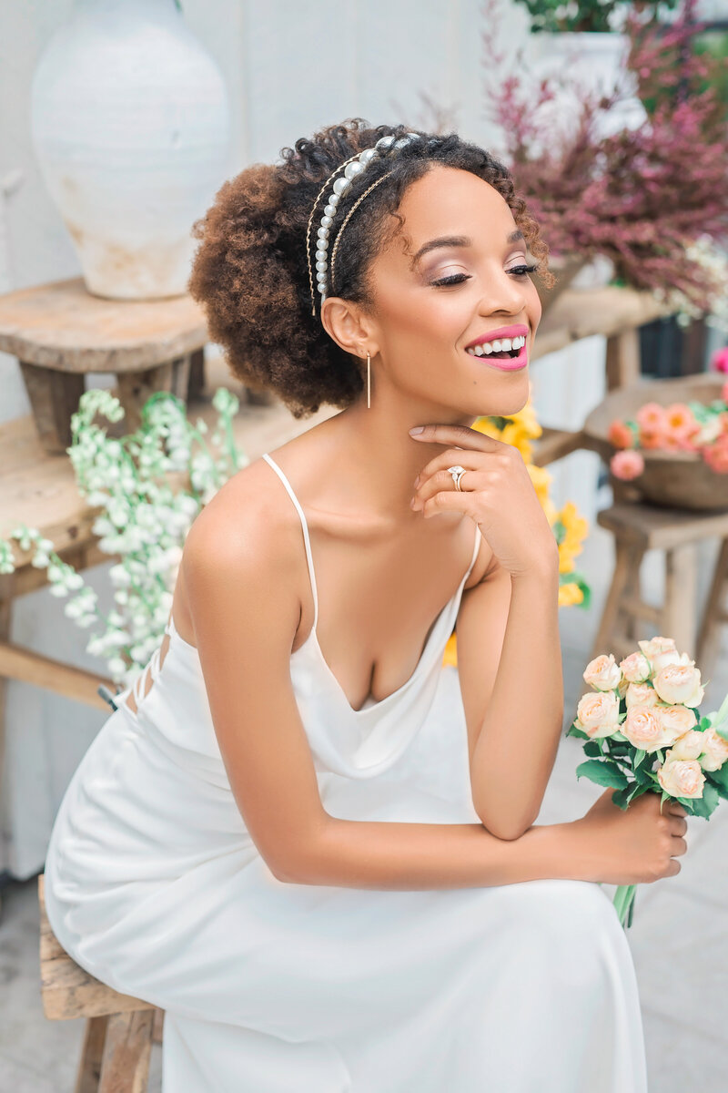A bride in a low-cut dress holds her chin and smiles to the distance. Behind here there are multiple bouquets.