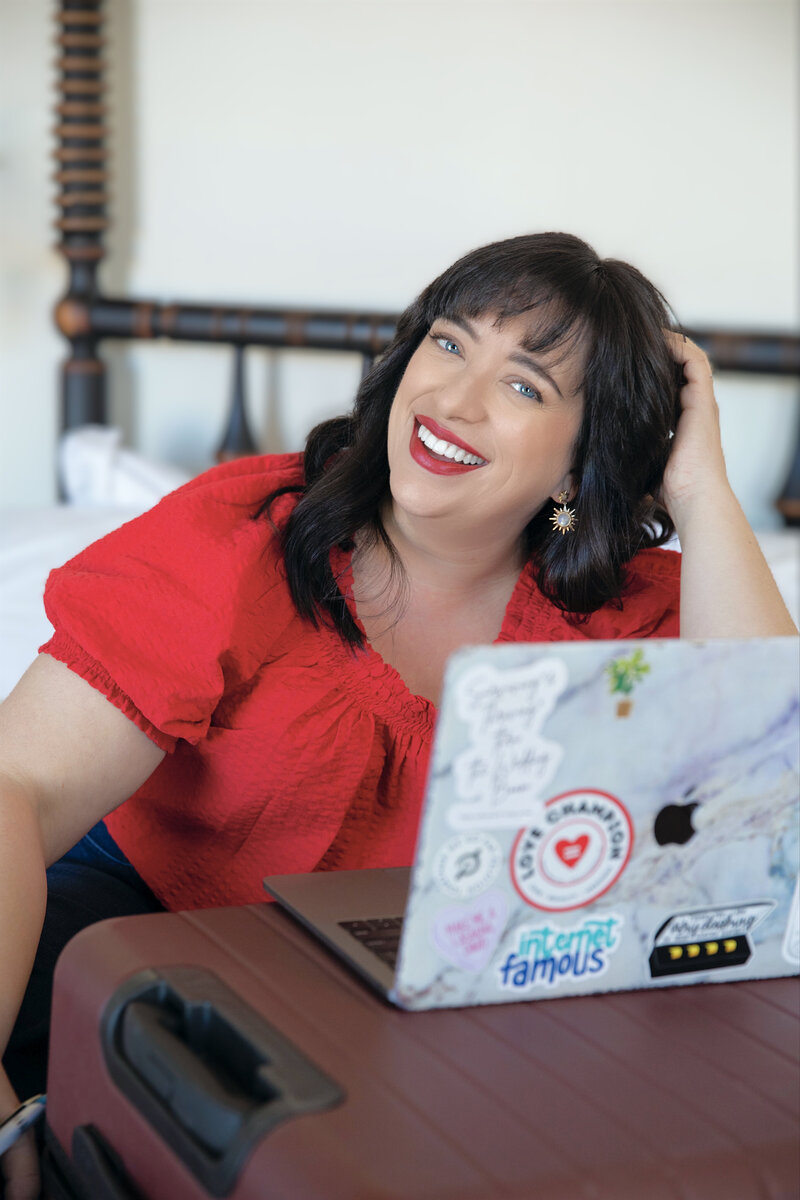Renee Dalo smiles in red blouse while sitting on hotel bed with her laptop