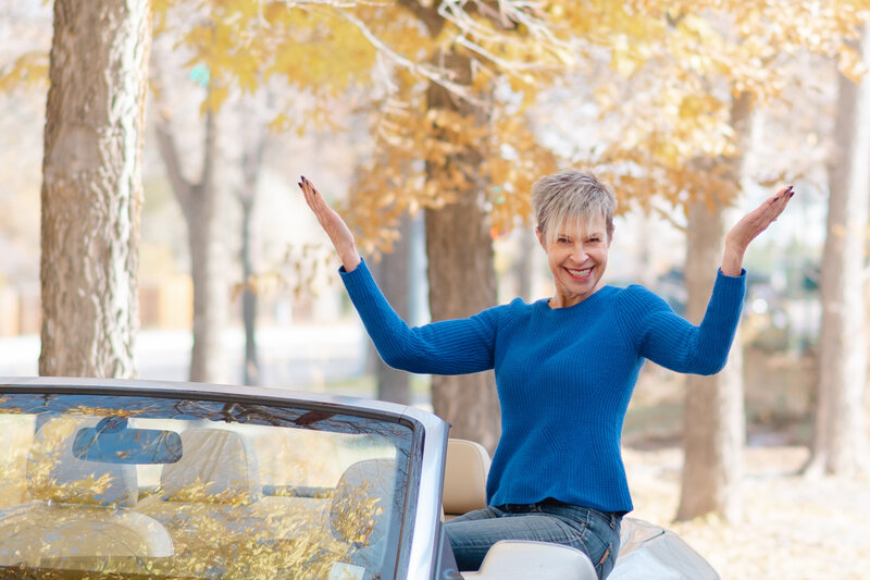 Lady sitting on side door of convertible with hands in air smiling with fall trees in background