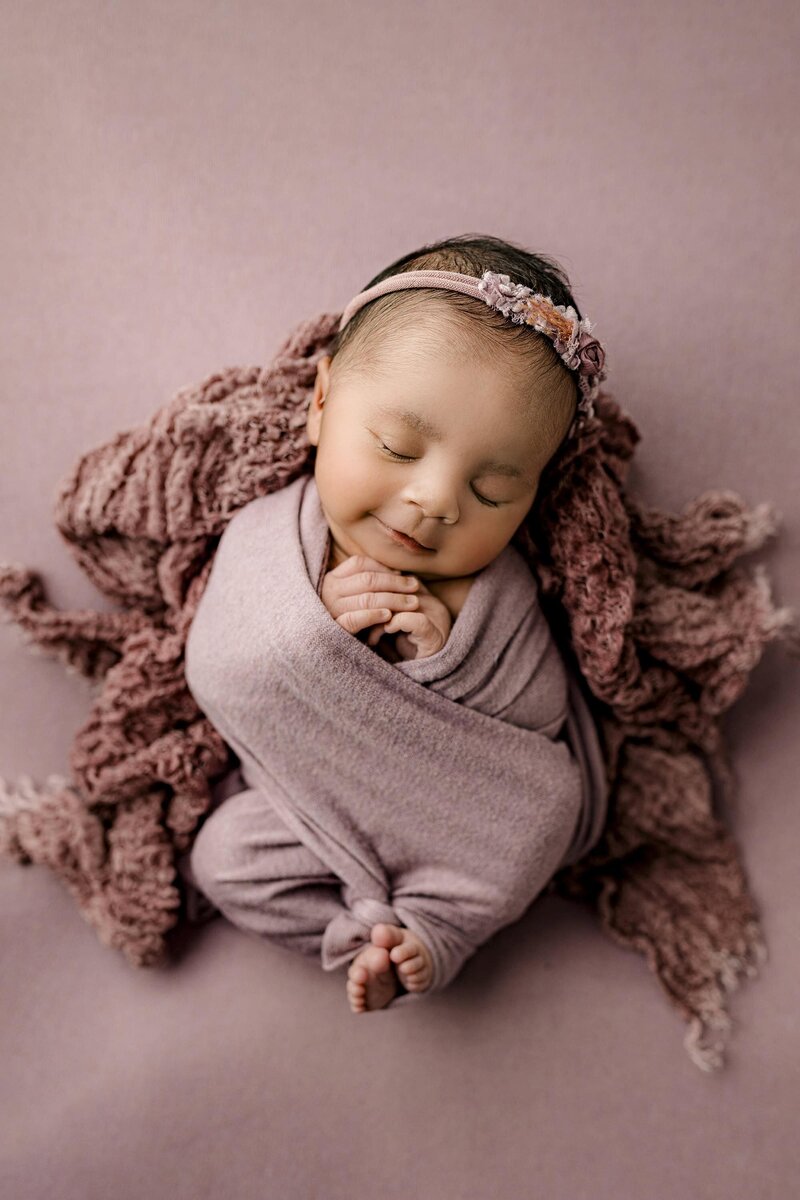 Newborn baby girl is wrapped in a lavender wrap with her toes peeking out in her hands underneath her chin her head tilted slightly to the side wearing a matching headband and on a purple layer background during their Studio newborn session