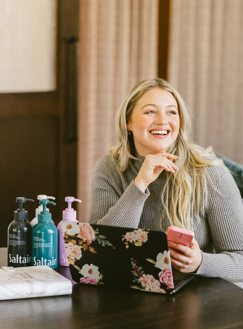 Iskra is sitting at a table with her laptop open and phone in hand. Her brands Saltair and Self Funding Planner are also on the table.