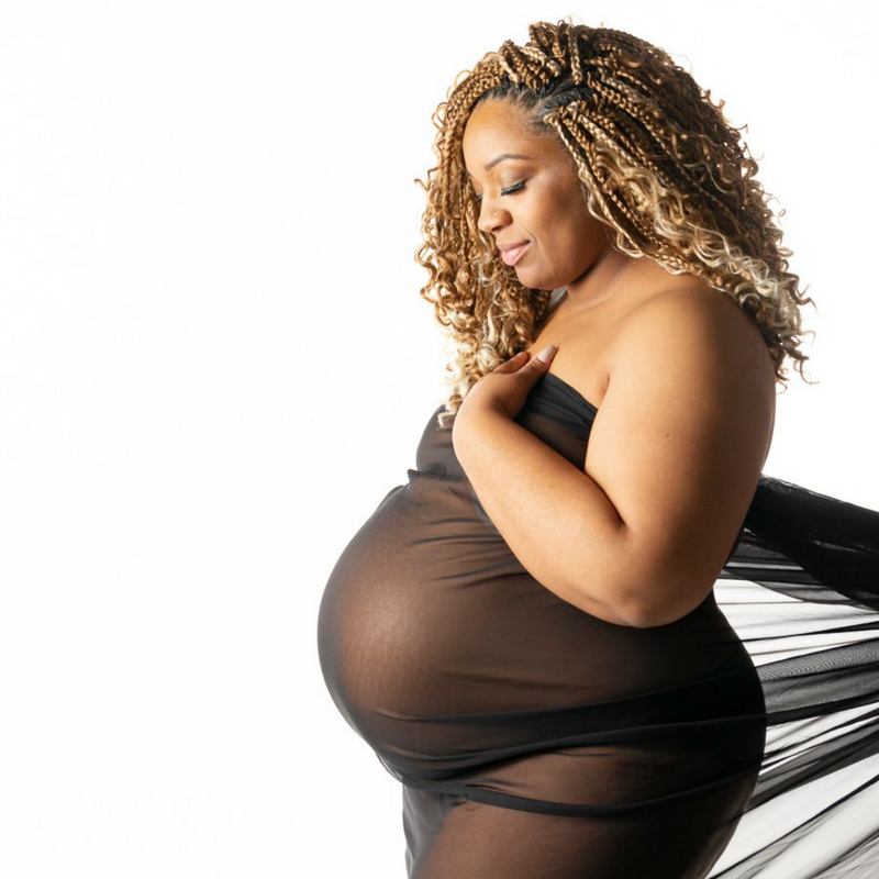 Dark skinned pregnant mom with blonde braids from Charlotte NC looking down at her pregnant belly draped in black fabric on a white background  created in our fort Mill portrait studio to celebrate her baby on the way