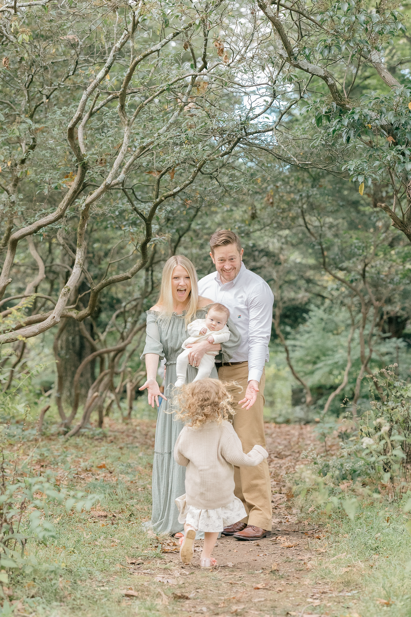 A mom and dad standing holding their newborn while they're older daughter runs towards them by NJ Family Photographer