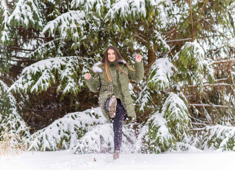 high school senior girl kicking snow with a snow covered pine tree behind her